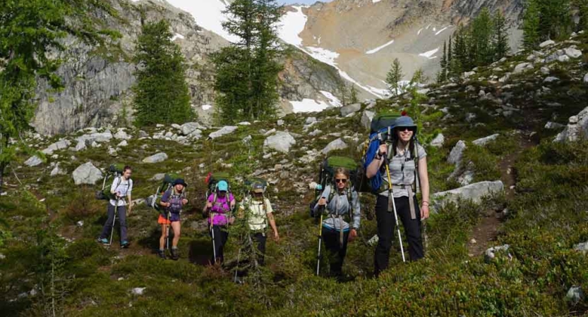 a group of students wearing backpacks and holding trekking poles hike along a grassy and rocky trail